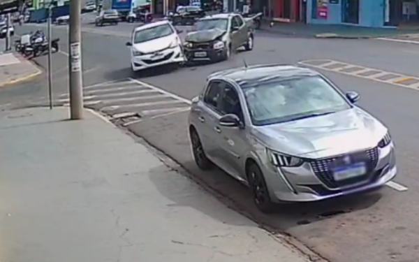 Imagem de uma câmera de segurança mostra o momento exato de uma colisão entre dois veículos em uma esquina urbana. Um carro branco (HB20) é atingido lateralmente por uma picape escura (Strada), que invade a preferencial vindo pela transversal. Ambos estão em movimento no instante da batida. Na frente da imagem, um terceiro carro, prata, aparece estacionado, e deve ter sido atingido na sequência do acidente. Ao fundo, é possível ver comércios, calçadas e outros veículos circulando.