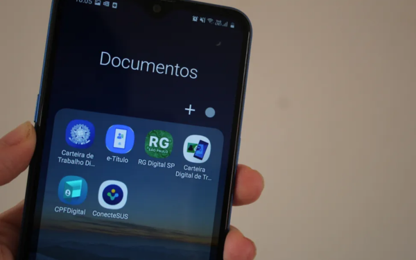 Pessoa segura um celular com a tela aberta na pasta “Documentos”. Nela, aparecem seis aplicativos do governo relacionados a documentos digitais: Carteira de Trabalho Digital, e-Título, RG Digital SP, Carteira Digital de Trânsito, CPF Digital e Conecte SUS. A imagem está bem iluminada, com fundo neutro.