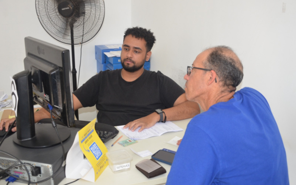 Na imagem, dois homens estão sentados em frente a um computador em um ambiente claro e simples. O homem à esquerda, mais jovem, veste camiseta preta e está concentrado na tela do computador enquanto manipula o mouse com a mão direita. O homem à direita, mais velho, usa óculos e camiseta azul, observa atentamente o monitor. Sobre a mesa há documentos, carteira, celular e uma placa amarela com QR code. Ao fundo, um ventilador de parede está ligado