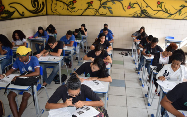 Sala de aula com vários estudantes do ensino médio sentados em carteiras enfileiradas. Eles estão concentrados preenchendo provas ou atividades impressas. A sala tem paredes azulejadas e um painel decorado na parte superior, com desenhos coloridos de flores e folhas.