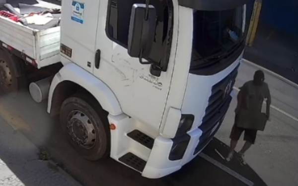 Imagem de uma câmera de segurança flagra o momento em que um homem, com boné, camiseta escura, bermuda e tênis, caminha ao lado de um caminhão branco estacionado na rua. O homem carrega uma bateria automotiva nos braços e parece deixar o local após o furto. A cena ocorre em plena luz do dia, e a sombra dos objetos indica forte iluminação solar