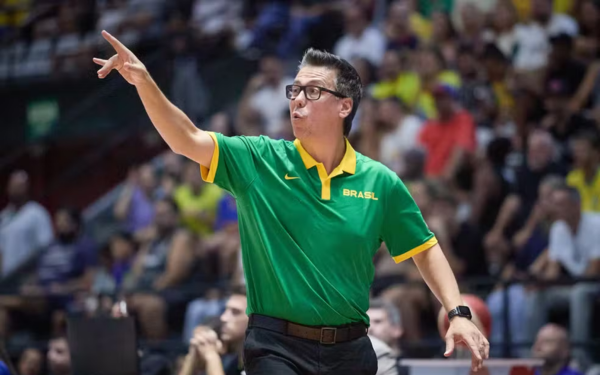 Homem de pele clara e cabelos escuros, usando óculos e uma camisa verde da Seleção Brasileira com detalhes amarelos e a palavra “BRASIL” no peito. Ele está em quadra, com o braço direito estendido e dois dedos apontando para frente, como se orientasse os jogadores. Ao fundo, há uma arquibancada cheia de torcedores, desfocados na imagem.