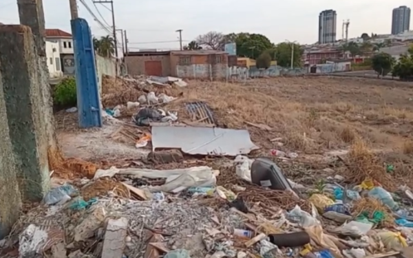 A imagem mostra um terreno baldio no centro de Franca, com o muro lateral quebrado e bastante lixo espalhado pelo chão. Há restos de entulho, sacolas plásticas, garrafas, pedaços de madeira e até um pneu velho. A área está tomada por mato seco, evidenciando abandono e falta de manutenção. Ao fundo, é possível ver casas, prédios e parte da cidade.