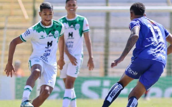 Na imagem, dois jogadores disputam a bola durante a partida entre Francana e Monte Azul. O atleta da Francana, de uniforme branco com detalhes em verde, conduz a bola pelo lado esquerdo do campo, enquanto o adversário do Monte Azul, de uniforme azul, se posiciona para a marcação. Ao fundo, outro jogador da Francana aparece sorrindo, acompanhando o lance.