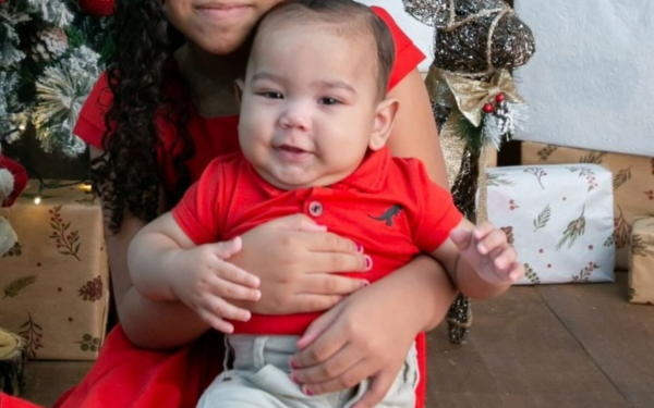 Um bebê de pele clara, cabelo curto e castanho, veste camisa polo vermelha com um pequeno desenho de dinossauro preto e calça clara. Ele está sorrindo e sentado no colo de uma criança mais velha, que usa vestido vermelho. Ao fundo, há uma árvore de Natal decorada, presentes embrulhados e um enfeite em formato de rena.