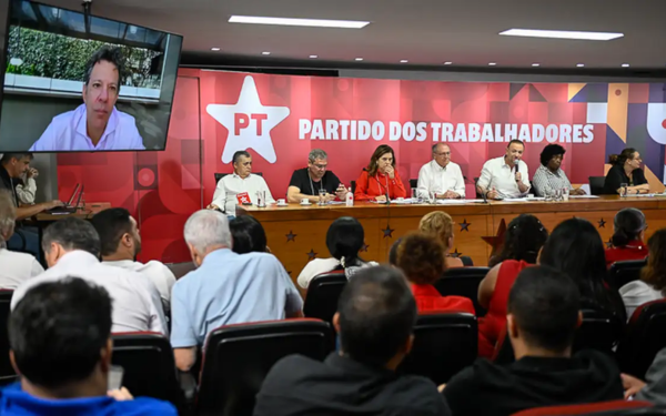 Auditório lotado com pessoas assistindo a um debate do Partido dos Trabalhadores (PT). Na mesa principal, vários integrantes do partido estão sentados lado a lado, incluindo parlamentares e lideranças, diante de um painel vermelho com o símbolo do PT. À esquerda, um telão exibe a imagem de um homem participando por vídeo.