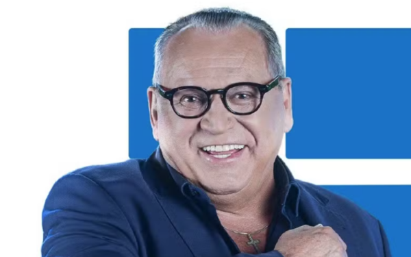 Homem de pele clara, usando óculos de aro preto, cabelo curto grisalho e penteado para trás. Ele veste um blazer azul escuro sobre uma camisa da mesma cor e sorri amplamente, com a mão direita apoiada no peito. Ao fundo, há formas geométricas azuis.