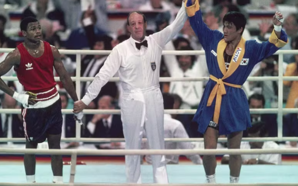 Na imagem, há uma cena de luta de boxe. No centro, um árbitro de camisa branca e gravata borboleta preta levanta o braço do lutador da direita, que veste um roupão azul com faixa amarela e traz a bandeira da Coreia do Sul no peito, indicando que ele é o vencedor. À esquerda, o outro boxeador, com uniforme vermelho e branco, aparece com expressão de descontentamento e o braço segurado pelo árbitro em sinal de derrota. Ao fundo, o público assiste ao momento.
