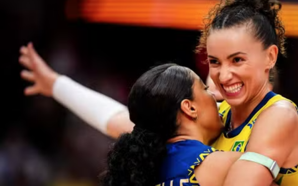 Duas jogadoras da seleção brasileira de vôlei comemoram um ponto durante a partida. Uma delas, vestindo uniforme amarelo com detalhes verdes, sorri amplamente enquanto abraça a companheira de equipe, que usa uniforme azul com o nome "MICHELLE" estampado nas costas. As duas transmitem emoção e alegria, com os braços erguidos em celebração.