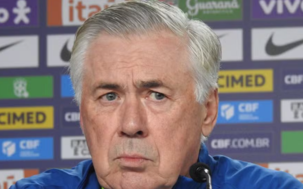Um homem de cabelos grisalhos curtos aparece em primeiro plano durante uma coletiva de imprensa. Ele veste uma jaqueta azul e olha com expressão séria. Ao fundo, há um painel com diversos logotipos de patrocinadores, incluindo Nike, Itaú, Guaraná Antarctica, Vivo e CBF.