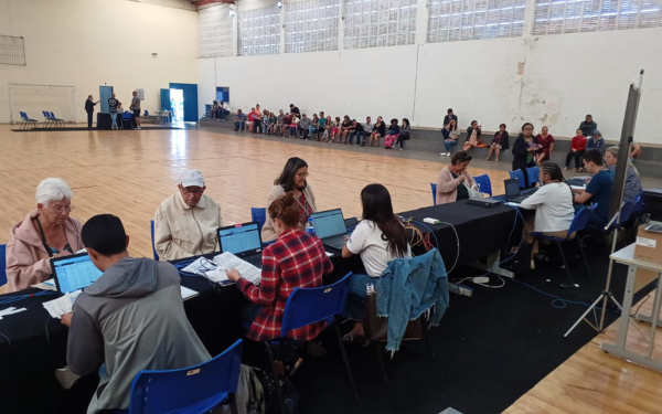 Dentro de um ginásio esportivo, várias mesas estão organizadas em fileira com atendentes da CDHU usando computadores. Pessoas, incluindo idosos e famílias, estão sentadas em frente aos atendentes para receber orientações. Ao fundo, outras pessoas aguardam sentadas em cadeiras na lateral da quadra.