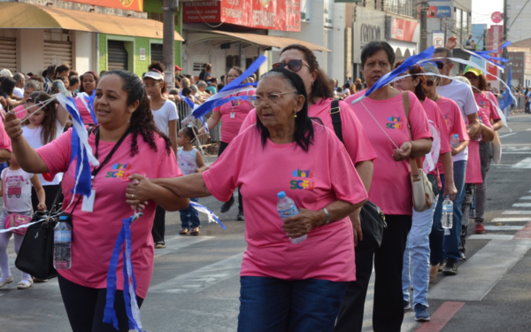 Um grupo de mulheres participa de um desfile cívico em uma rua movimentada. Elas vestem camisetas cor-de-rosa com o logotipo da Secretaria de Educação e carregam fitas azuis nas mãos, que balançam enquanto caminham. Algumas seguram garrafas de água. Ao fundo, mais pessoas acompanham o desfile, incluindo crianças e famílias nas calçadas.