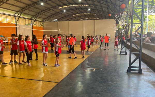 Dentro de um ginásio coberto, dezenas de crianças vestindo camisetas vermelhas participam de atividades de basquete. Algumas estão em fila aguardando a vez, enquanto outras praticam arremessos em tabelas posicionadas no espaço. Professores acompanham a movimentação e, nas arquibancadas ao lado, algumas pessoas assistem.