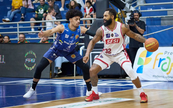 Jogador do Franca Basquete, com uniforme branco e número 32, segura a bola com a mão direita enquanto é marcado de perto por atleta do São José, que veste uniforme azul. A cena acontece dentro de quadra, com torcedores e equipe de transmissão ao fundo.