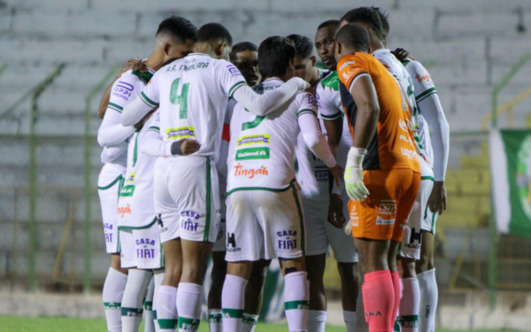 Jogadores da Francana, vestidos com o uniforme branco com detalhes em verde, estão reunidos em roda dentro de campo, abraçados em concentração antes do início da partida. O goleiro, de uniforme laranja, também participa do momento. Ao fundo, arquibancadas vazias do estádio.