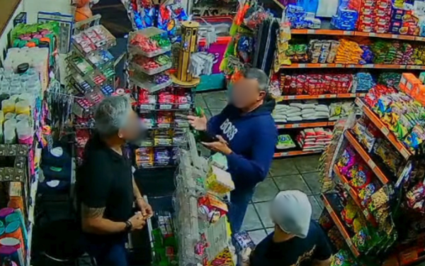 Imagem de câmera de segurança dentro de um mercado mostra três pessoas próximas ao caixa. À esquerda, um homem de camiseta preta, possivelmente o dono do estabelecimento, está em pé diante do balcão. No centro, outro homem de moletom azul faz gestos com as mãos, aparentando discutir. À frente, um terceiro cliente, de boné branco, observa a cena. As prateleiras ao redor estão cheias de produtos, como pacotes de biscoitos e doces.