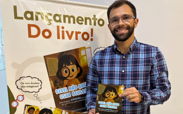 Homem de barba, óculos e camisa xadrez azul segura um exemplar do livro infantil “Bebel Não Quer Usar Óculos” em frente a um banner de divulgação do lançamento da obra. No cartaz, aparece a ilustração da personagem principal da história, uma menina de óculos com olhar alegre.
