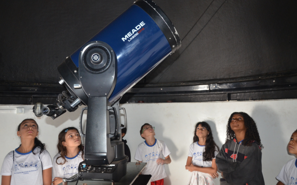 Um grupo de crianças e adolescentes observa com curiosidade um grande telescópio azul, instalado em uma sala do observatório. Elas olham para cima, demonstrando atenção e interesse pelo equipamento e pelo céu.