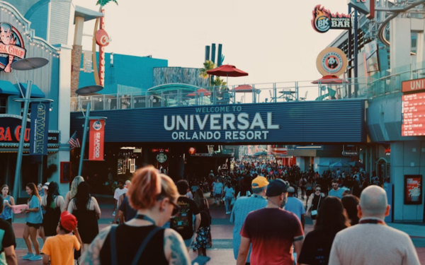 Foto mostra a entrada do Universal Orlando Resort, com um grande letreiro azul que dá boas-vindas aos visitantes. A cena está repleta de pessoas caminhando em direção ao parque, algumas em grupos, outras sozinhas, em clima de lazer. Ao fundo, é possível ver prédios temáticos, restaurantes e letreiros luminosos que compõem o ambiente vibrante do complexo.