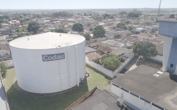 A imagem mostra um grande reservatório de água cilíndrico, pintado de branco, com a logomarca da Codau em destaque. Ele está localizado em uma área urbana, cercado por casas, árvores e algumas construções. Ao fundo, é possível ver a cidade se estendendo até o horizonte.