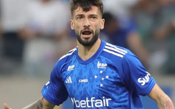 A foto mostra um jogador de futebol em campo, usando o uniforme azul do Cruzeiro. Ele aparece em movimento, com a boca entreaberta, como se estivesse falando ou comemorando. A camisa tem detalhes brancos e o patrocínio da Betfair estampado na parte da frente. Ao fundo, vê-se a torcida desfocada nas arquibancadas.