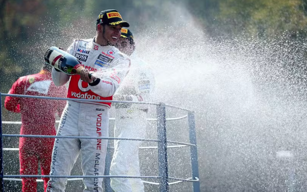 Na imagem, um piloto de Fórmula 1 comemora no pódio. Ele veste macacão branco com detalhes vermelhos da equipe Vodafone McLaren Mercedes e boné preto com a logo da Pirelli. O piloto segura uma garrafa de champanhe e a agita, espalhando jatos de espuma para o alto e para os lados. Ao fundo, outros competidores também participam da celebração, parcialmente borrados pelo spray da bebida. A cena transmite alegria e vitória.