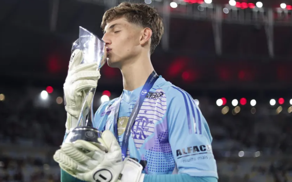 Um jovem goleiro, vestindo uniforme azul claro e luvas brancas, aparece na imagem beijando um troféu prateado em campo. Ele usa uma medalha pendurada no pescoço e está em um estádio iluminado, com arquibancadas ao fundo.
