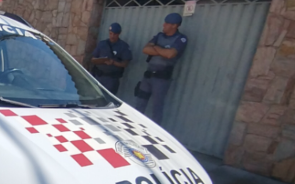 A imagem mostra parte de uma viatura da Polícia Militar estacionada em frente a um portão. Dois policiais de uniforme azul estão ao lado do portão cinza: um deles escreve em uma prancheta, enquanto o outro permanece de braços cruzados, observando o local. A cena sugere atuação policial em uma ocorrência.