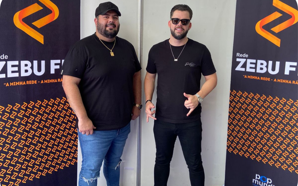 Na foto, dois homens posam sorridentes em frente a dois painéis da rádio Zebu FM. Ambos vestem camiseta preta e calça jeans. O homem à esquerda usa boné preto, corrente dourada e relógio, enquanto o da direita usa óculos escuros e corrente prateada, fazendo um gesto com as mãos.