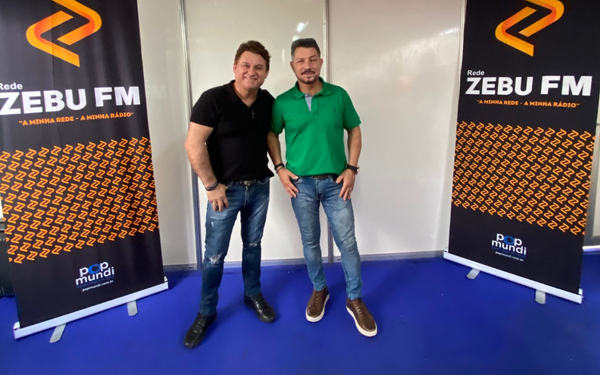 Na foto, dois homens posam lado a lado em frente a dois painéis da rádio Zebu FM. O homem à esquerda veste camiseta preta e calça jeans, enquanto o da direita usa camisa polo verde e também calça jeans. Ambos sorriem e estão em um ambiente com piso azul.