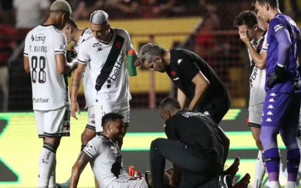 Jogador do Vasco está sentado no gramado, recebendo atendimento médico durante a partida. Ao redor dele, companheiros de equipe e membros da comissão técnica observam a situação com preocupação