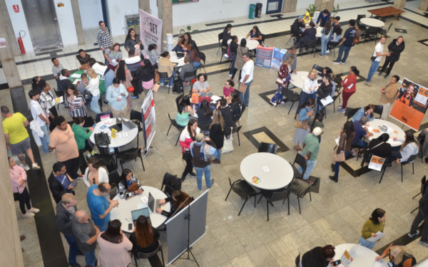 Fotografia mostra um grande espaço interno com várias mesas distribuídas, onde pessoas conversam e entregam currículos. O ambiente está cheio de candidatos e representantes de empresas, alguns sentados e outros em pé, formando filas. Há cartazes e banners próximos às mesas, indicando os diferentes setores de contratação. A cena retrata um evento de recrutamento, com movimentação intensa de pessoas em busca de oportunidades de trabalho.