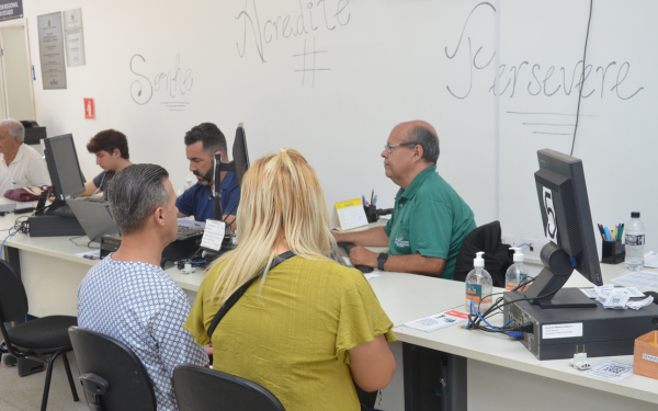 Em uma sala de atendimento, pessoas estão sentadas de frente para atendentes em mesas com computadores. Um homem de camisa verde conversa com um casal, enquanto outros candidatos aguardam orientação em guichês ao lado. Na parede ao fundo, palavras motivacionais como “Sorria”, “Acredite” e “Persevere” estão escritas em letras grandes.
