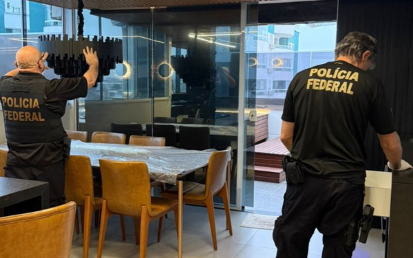 Dois policiais federais, de costas, realizam busca em um apartamento moderno. Um deles examina uma luminária sobre a mesa de jantar e o outro abre uma gaveta. Ambos vestem camiseta preta com a inscrição “Polícia Federal”. O ambiente tem cadeiras de couro, mesa de vidro e vista para prédios pela sacada envidraçada.