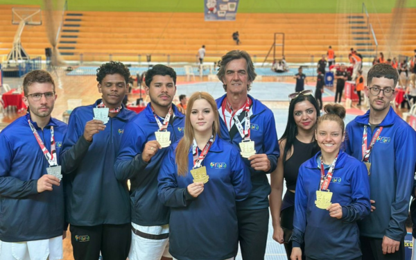 Grupo de atletas de taekwondo posa em quadra esportiva segurando medalhas conquistadas na Copa São Paulo. São seis atletas jovens, todos uniformizados com jaquetas azuis, acompanhados por um treinador e uma mulher ao lado, em clima de comemoração. Ao fundo, vê-se arquibancadas e parte da estrutura do ginásio.