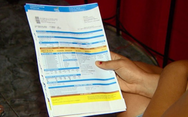 Uma pessoa segura uma conta de energia elétrica aberta com as duas mãos. O documento tem várias linhas em azul e branco, com informações detalhadas sobre consumo e valores cobrados. A mão que segura a conta está com as unhas pintadas de vermelho.
