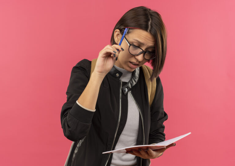 Thoughtful young student girl wearing glasses and back bag holding and looking at note pad touching head with pen isolated on pink background with copy space